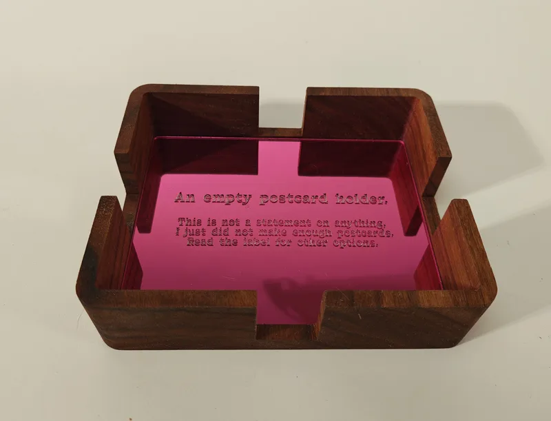 The empty walnut holder revealing its mirror-pink acrylic base, engraved: An empty postcard holder. This is not a statement on anything, I just did not make enough postcards. Read the label for other options.
