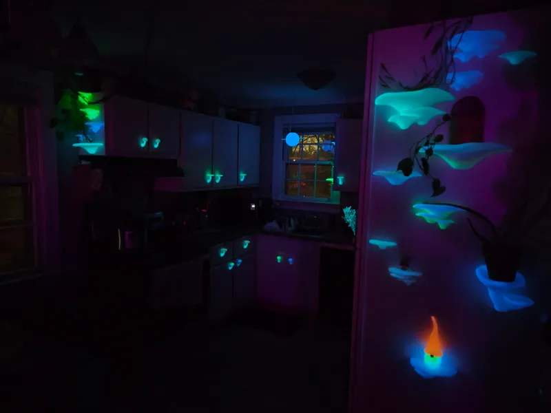 The full installation glowing after dark: blue and green mushroom caps climbing the cabinets, walls, and refrigerator across a darkened kitchen