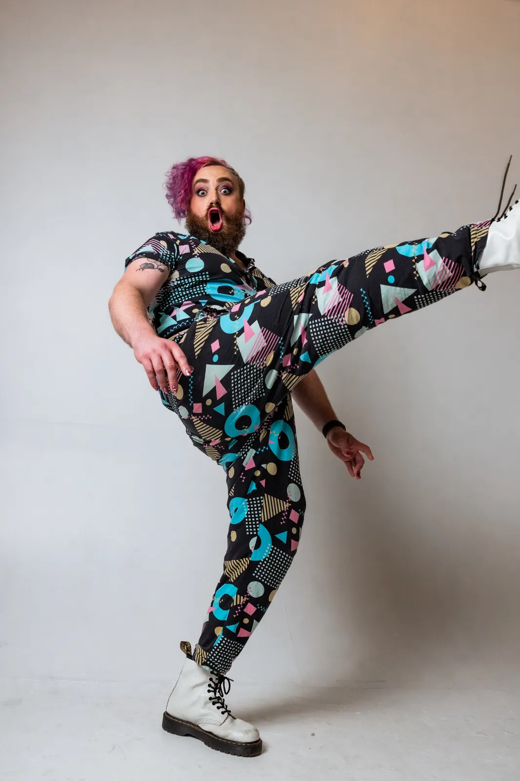 Kev mid-kick, pink hair and beard, geometric-print jumpsuit, white Doc Martens, in a studio
